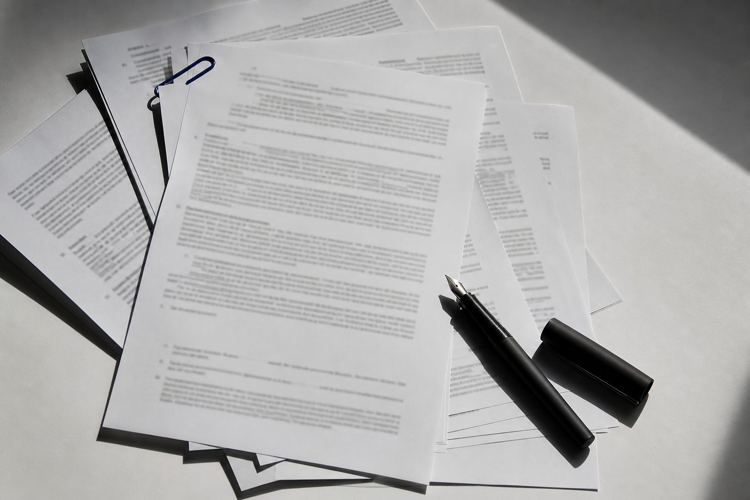 Printed contract pages fanned on a white desk with a black fountain pen and an indigo paper clip