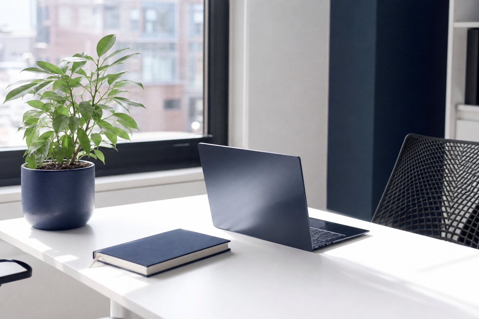 Bright minimalist office desk with an open laptop, a notebook and a small plant beside a large window